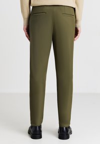 Olivgrüne, maßgeschneiderte Hose mit glatter Textur. Zwei dezent gestaltete Gesäßtaschen, kombiniert mit schwarzen Schuhen für den Kontrast.