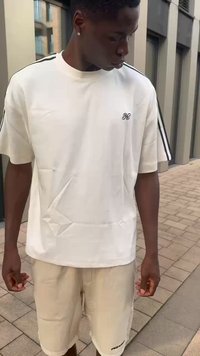 Weißes Baumwoll-T-Shirt mit kurzen Ärmeln und seitlichen Streifen; verfügt über ein kleines gesticktes Logo. Kombiniert mit beigen Shorts und Sneakers.