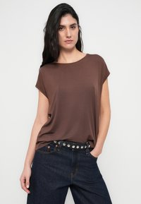 Καφέ κοντομάνικη μπλούζα με χαλαρή εφαρμογή και στρογγυλή λαιμόκοψη, φορεμένη με σκούρο τζιν και μαύρη ζώνη με καρφιά. Λεία υφή.