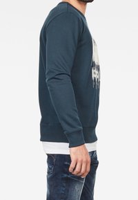 Man som bär en mörkblå sweatshirt med ett vitt grafiskt tryck, lager på en vit skjorta, och blekta blå jeans, visad från sidan.