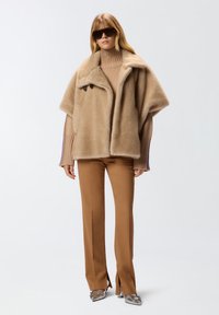 Donna con un ampio cappotto in pelliccia sintetica beige, un maglione a collo alto beige, pantaloni marroni con spacchi frontali, tacchi a punta argentati e occhiali da sole oversize neri.