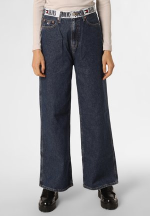 Osoba ubrana w ciemnoniebieskie szerokie jeansy, biały pasek z logo Tommy Jeans, beżową bluzkę z długim rękawem i czarne masywne buty.