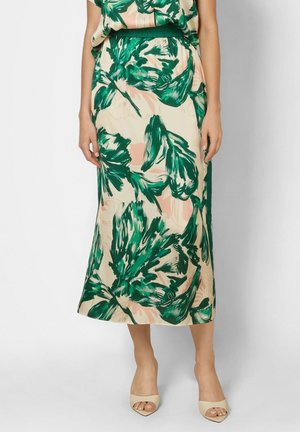 Woman wearing beige midi skirt with green and light pink abstract leaf pattern, paired with beige open-toe heels, standing on a plain background.