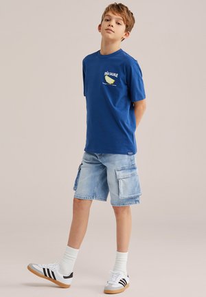 Poiss seisab käed selja taga, seljas sinise graafilise kujundusega T-särk, heledad teksakangast cargo-püksid, valged sokid ja valged tossud mustade triipudega.