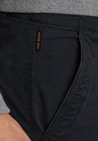 Schwarze lässige Hosen aus glattem Stoff mit Seitentasche, sichtbaren Nähten und einem goldenen Logosticker "PALE LEGEND."