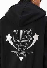 Svart hettegenser med hvit brodert tekst "GUESS" og "FOLLOW YOUR DESTINY" med stjerne- og lynbolt-grafikk. 
