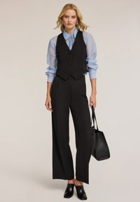 Schwarze Weste mit Knöpfen, hellbluse durchsichtige Langarmbluse, schwarze weit geschnittene Hose und schwarze Mules. Kombiniert mit einer schwarzen Handtasche.