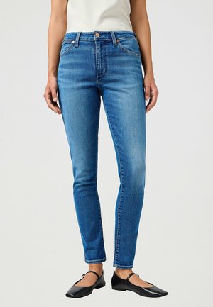 Femme portant un jean skinny bleu, un haut blanc et des chaussures plates noires avec brides à la cheville, se tenant devant un fond clair uni.