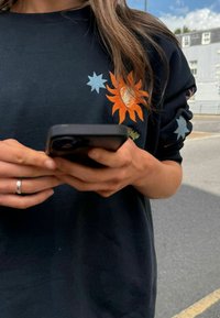 Czarna bluza z kolorowymi haftowanymi wzorami słońca i gwiazdy. Osoba trzymająca smartfon, pokazująca gołe dłonie z pierścionkiem.