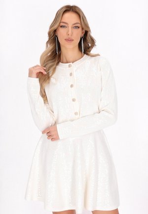 Blonde vrouw die een witte lange mouwen paillettenjurk draagt met knopen aan de voorkant en lange hangende oorbellen.