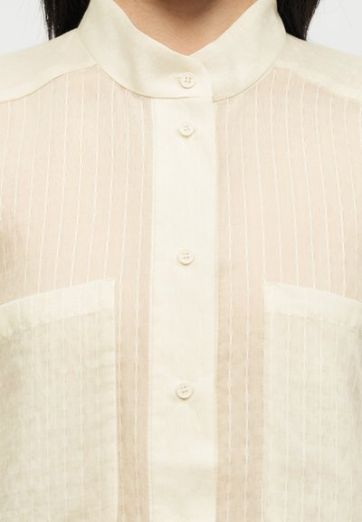 Blouse couleur crème avec des rayures verticales texturées, boutonnée à l'avant, col montant et deux poches poitrine.