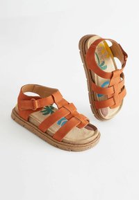 Bruna mockaskinnsandaler med justerbara remmar, en vadderad innersula med ett färgglatt tropiskt mönster och en texturerad gummisula.
