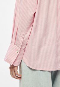 Camisa rosa com riscas finas verticais, com punho abotoado e nome da marca bordado. O tecido parece macio e com textura suave.