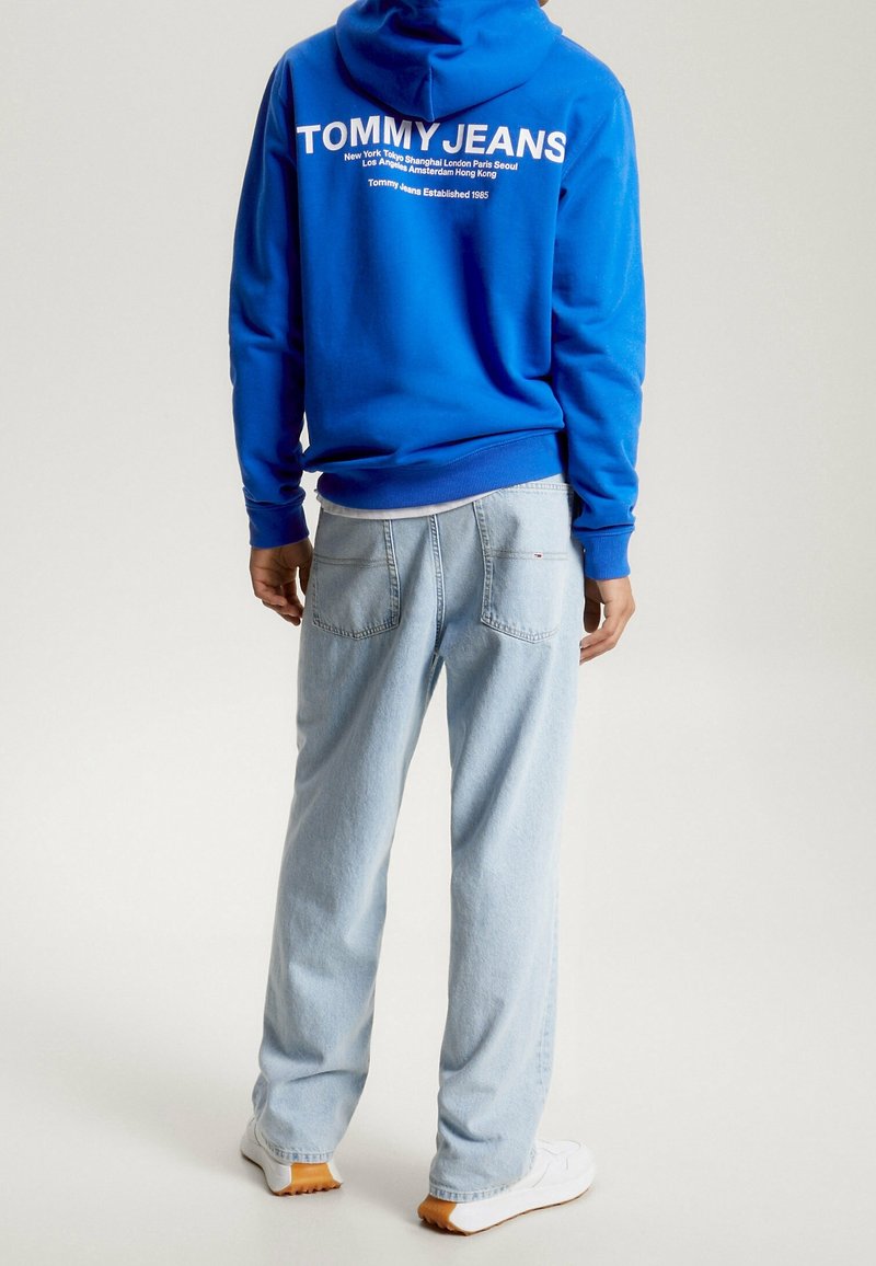 Sweat à capuche bleu avec texte blanc au dos, assorti à un jean en denim bleu clair. Baskets blanches ; coupe décontractée avec poches arrière visibles.