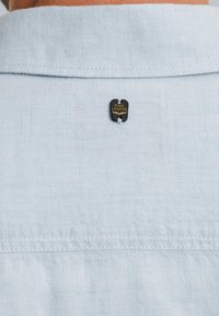 Cuello de camisa de lino azul claro con una etiqueta PME Legend en negro y dorado, que presenta costuras blancas a lo largo de la costura y una tela suave y texturizada.