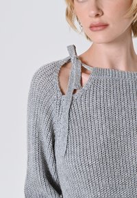 Primo piano di una donna che indossa un maglione lavorato a maglia grigio chiaro con taglio sulla spalla e dettaglio del fiocco annodato.