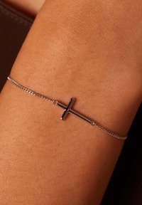 Pulsera de cadena de plata con un simple encanto de cruz. La cadena es delicada y la cruz tiene un acabado pulido con formas rectangulares.