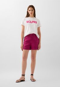 Bílá tričko s růžovým potiskem "SCLPRS", doplněné jasně magenta šortkami. Model nosí černé sandály na pásky se jednoduchým designem a plochým profilem.