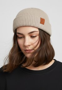Beige knitted beanie with a folded ribbed cuff and a rectangular leather patch. The fabric has a soft texture, complementing a black top.