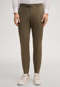 Mann trägt schlanke olivgrüne Hose, weiße Sneaker und ein langärmliges weißes Hemd, steht vor einem einfarbigen Hintergrund.