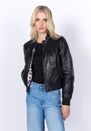 Blondýna v černé kožené bundě s leopardím potiskem na podšívce, černém tričku a světle modrých džínách na jednobarevném bílém pozadí.