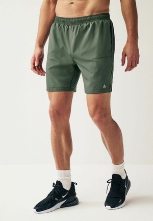 Pantaloni sportivi verdi con cintura elastica, tasche laterali e dettaglio triangolare del logo. Indossati con scarpe da ginnastica nere e calze bianche.