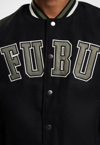 Sorte varsityjakke med stort broderet "FUBU"-skrift i grå og hvid, med sølvknapper og en stribet krave.