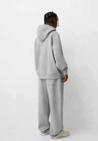 Sudadera gris con capucha de ajuste holgado y hombros caídos, combinada con pantalones deportivos a juego. Textura suave, diseño casual y silueta sobredimensionada.