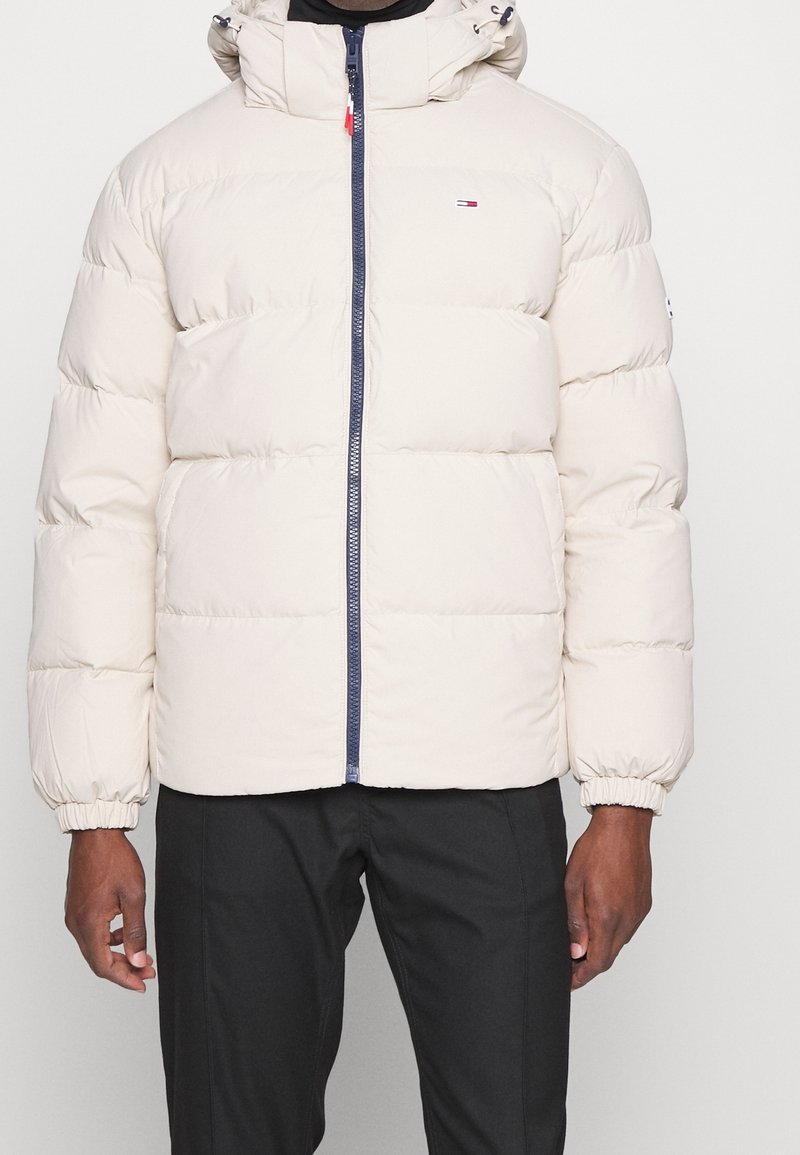 Homme portant une doudoune beige matelassée avec une capuche et une fermeture éclair bleu marine, associée à un pantalon noir, debout devant un fond uni.