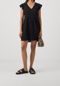 Svart kort klänning med V-hals, volangärmar och texturerat tyg. Matchas med svarta sandaler med remmar och en liten beige handväska.
