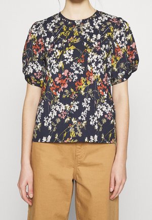 Femme portant un chemisier bleu marine à imprimé floral blanc, rouge et jaune avec des manches courtes bouffantes, associé à un pantalon marron clair.