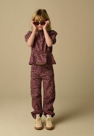 Child in matching purple leopard-print top and pants adjusting dark sunglasses against beige background.