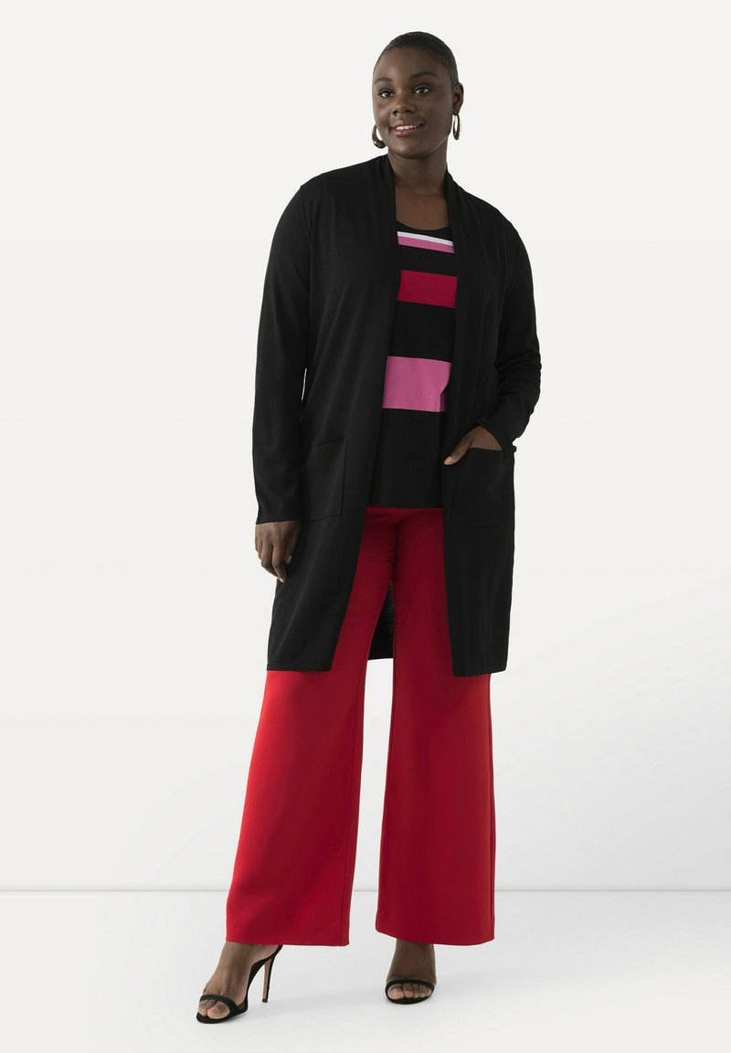 Schwarzer langer Cardigan mit zwei vorderen Taschen, über einem bunten gestreiften Oberteil in Pink, Rot und Schwarz getragen, kombiniert mit weiten roten Hosen.