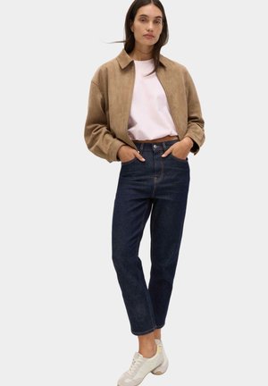 Kvinde iført tanfarvet ruskindjakke, lyserød skjorte, mørkeblå jeans og hvide sneakers, stående med hænderne i lommerne mod hvid baggrund.
