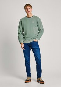Grön sweatshirt med rund halsringning och logodetalj, i kombination med blå jeans och flerfärgade sneakers, ståendes mot en neutral bakgrund.