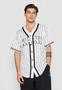 Vit baseballtröja med svarta vertikala ränder, "KANI"-logotyp på framsidan, V-hals, kortärmad, knappar framtill, ihopparad med svarta byxor.