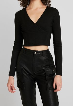 A woman models a black ribbed crop top with a V-neck, paired with high-waisted black leather cargo pants, against a plain background.