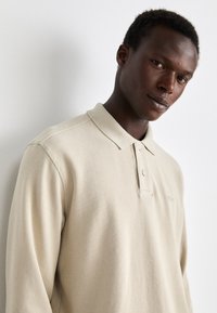 Polo de manga longa bege e canelado com colarinho, apresentando uma pala com botões e um detalhe discreto do logótipo, apresentado contra um fundo neutro.