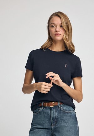 Dunkelblaue Baumwoll-T-Shirt mit kurzen Ärmeln, rundem Kragen, versehen mit einem kleinen gestickten Logo auf der Brust und kombiniert mit hochgeschnittenen Jeans.