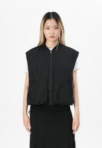 Vrouw in een mouwloze zwarte gewatteerde vest met een rits en knopen aan de voorkant, over een witte blouse en zwarte rok, staand voor een witte achtergrond.