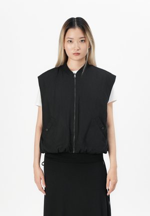 Woman wearing a sleeveless black padded vest with front zipper and buttons over a white shirt and black skirt, standing against white background.