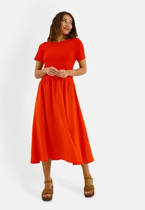 Mujer de pie con las manos juntas, vestida con un vestido corto de manga corta de color naranja brillante y sandalias marrones de plataforma, mirando de lado con una sonrisa.