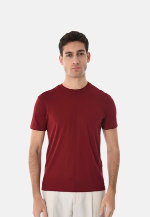 Giovane uomo in piedi di fronte, indossa una maglietta a maniche corte rossa semplice e pantaloni chiari su uno sfondo bianco.