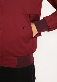 Giacca bordeaux con chiusura a bottoni, polsini a coste e tasche laterali. Realizzata in tessuto liscio con una fascia in vita nera a contrasto.