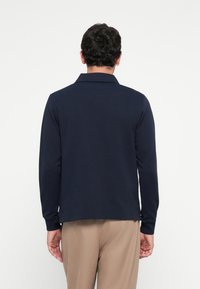 Man standing facing away wearing a navy long-sleeve polo shirt and beige pants against a plain white background.