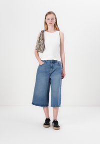 Bílé bezrukávové tričko, modré široké džínové culotky a černé tenisky. Modelka nese kabelku s texturou hadího vzoru. Jednoduchý a ležérní outfit.