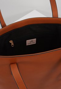 Hnědá kožená taška přes rameno s černým interiérem, viditelnou zipovou kapsou a štítkem "Anna Field" uvnitř.