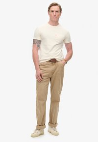 Creme t-shirt med lomme, parret med beige chinos. Modellen bærer et brunt bælte og hvide sneakers. Glat tekstur hele vejen igennem.