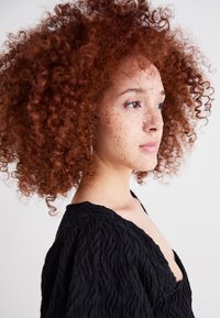 Capelli ricci rossi, pelle con lentiggini e un indumento nero strutturato con un profondo scollo a V. Vista laterale, sfondo neutro.