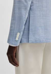 Ljusblå linnejacka med en texturerad väv, utrustad med en enkel vent och fyra vita knappar på manschetten. Tillsammans med beige byxor.